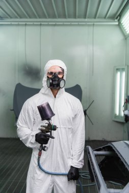 Koruyucu üniformalı genç bir işçi aerograf tutuyor ve otoparkta arabanın yanındaki kameraya bakıyor. 