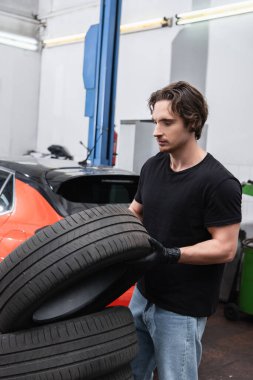 Young workman holding tire while working in car service  clipart