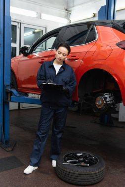 Mechanic in uniform writing on clipboard near tire, screwdriver and car in garage  clipart