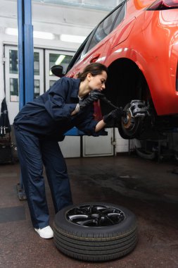 Side view of mechanic working with screwdriver and car wheel in garage  clipart