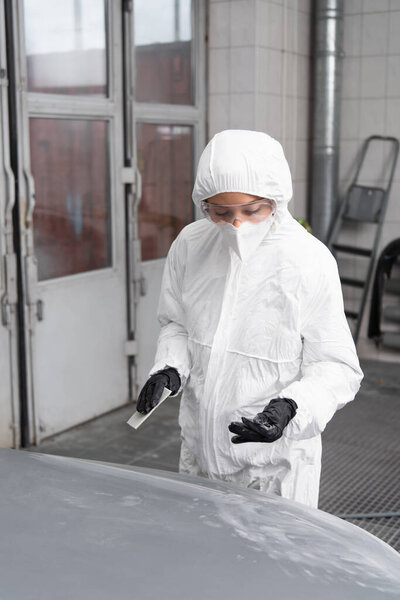 Workwoman in protective suit and mask holding sandpaper near car in service 