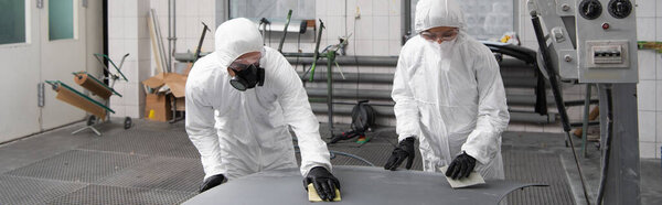 Mechanics in protective suits working with sandpaper and car hood in garage, banner 