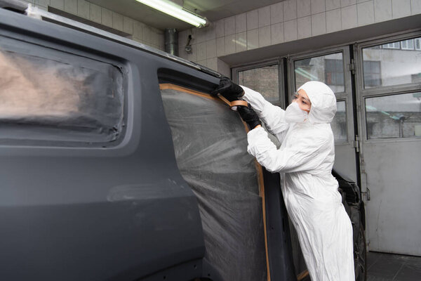 Workwoman in hazmat suit applying tape on car in service 
