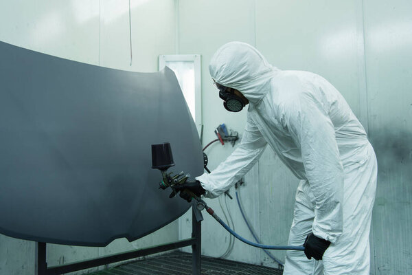 Workman in protective suit spraying varnish on car part in garage 