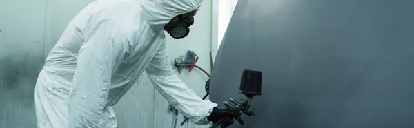 Workman in hazmat suit spraying varnish on car part in garage, banner