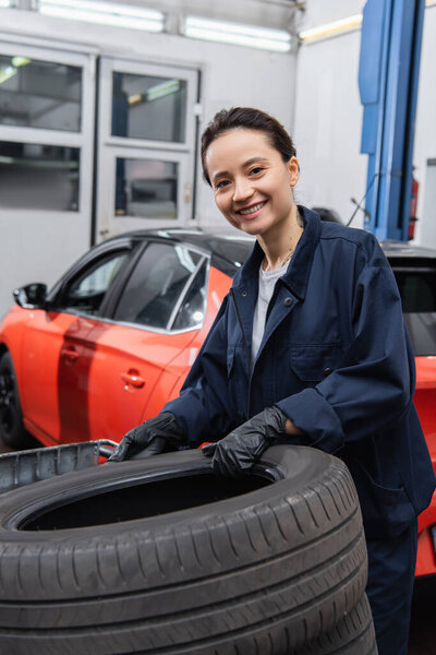 Mechanic in gloves and uniform smiling at camera near tires and car in garage 
