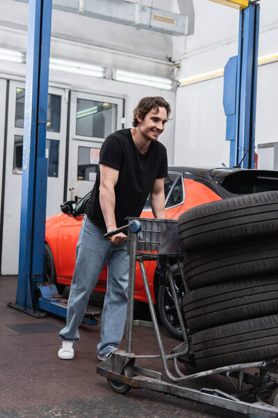 Cheerful mechanic standing near car with tires and auto in garage 