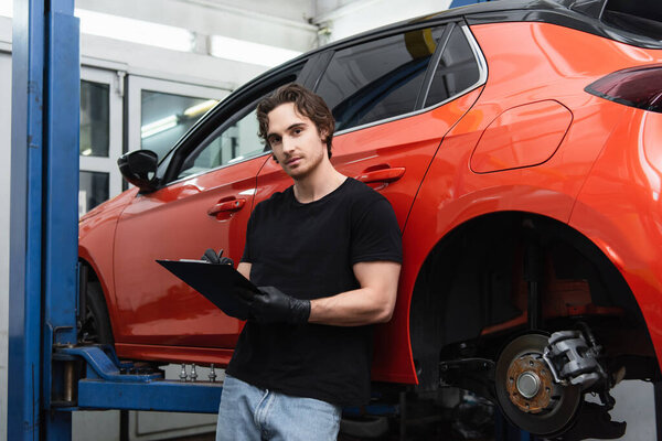 Young mechanic writing on clipboard and looking at camera near car in garage 
