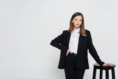 young stylish woman in suit posing with hand on hip near chair isolated on grey