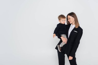 stylish and happy mother holding in arms toddler son isolated on grey