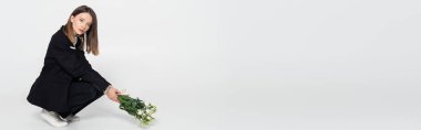 trendy young woman in black suit sitting and holding white flowers on grey, banner
