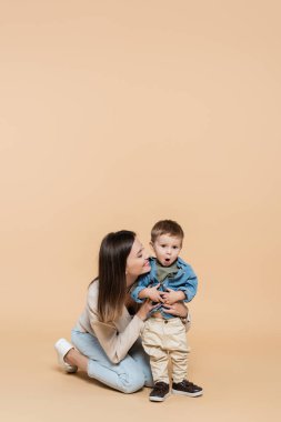 happy mother hugging surprised toddler son on beige