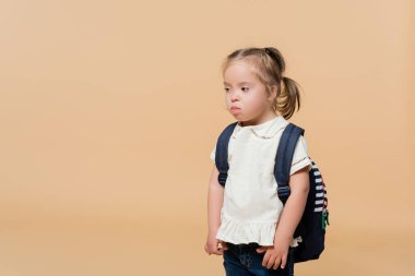 girl with down syndrome sticking out tongue while standing with backpack on beige 