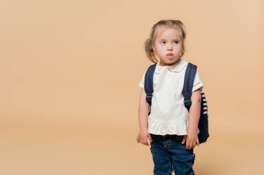 kid with down syndrome sticking out tongue while standing with backpack on beige 