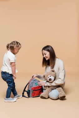 happy mother holding teddy bear and backpack near girl with down syndrome on beige