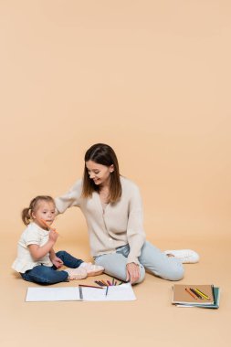 kid with down syndrome sitting with happy mother near colorful pencils on beige