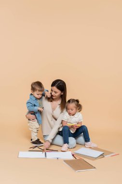 cheerful mother sitting near girl with down syndrome, toddler boy and colorful pencils on beige