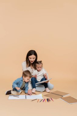 happy mother sitting near girl with down syndrome and toddler boy drawing on beige