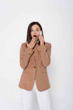 shocked young woman in beige blazer talking on smartphone and covering open mouth on white