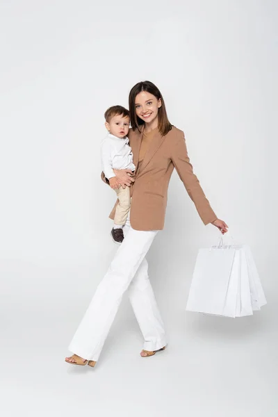 腕の中で楽しいお母さん幼児の息子と買い物袋を持ってグレーを歩きながら — ストック写真