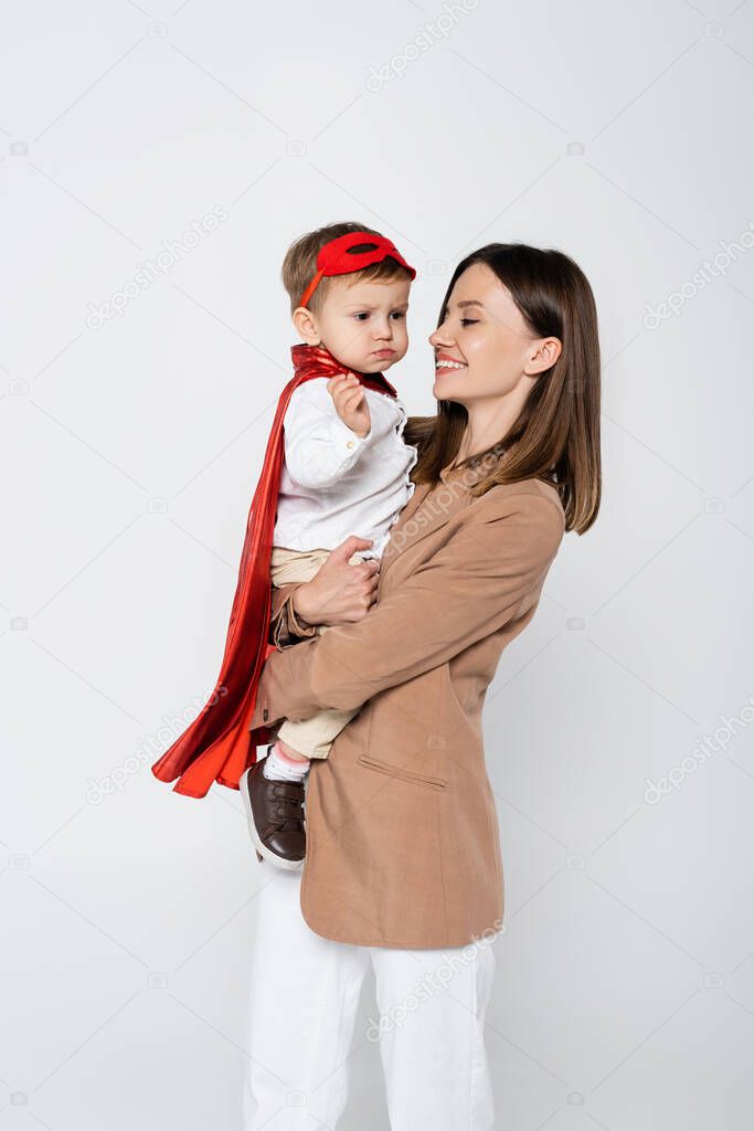 happy mother holding toddler son in superhero costume isolated on grey