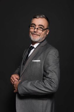 joyful man in blazer and eyeglasses looking at camera isolated on dark grey