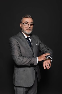 senior businessman in blazer, eyeglasses and wristwatch looking at camera on dark grey