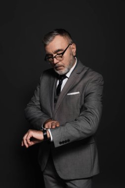 senior man in eyeglasses and formal wear looking at wristwatch on dark grey