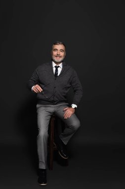 full length view of senior man in stylish clothes sitting on high stool and smiling at camera on grey