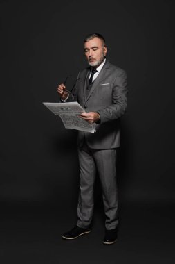 full length view of elderly man in formal wear holding eyeglasses while reading newspaper on dark grey