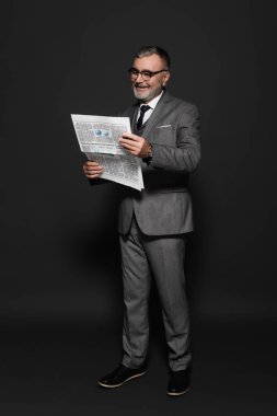 full length of senior man in suit and eyeglasses reading newspaper on dark grey