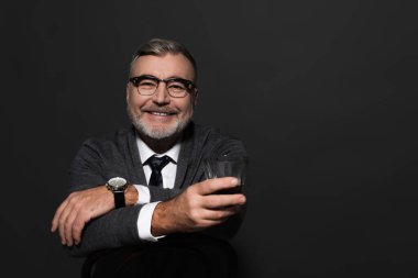 happy senior man in stylish clothes holding glass of whiskey and looking at camera while sitting isolated on dark grey
