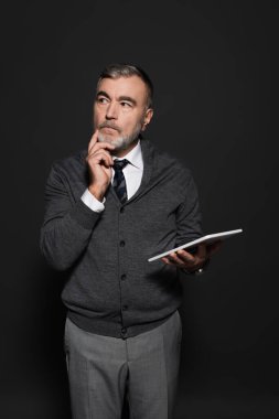 pensive man holding digital tablet and hand near face while looking away on dark grey
