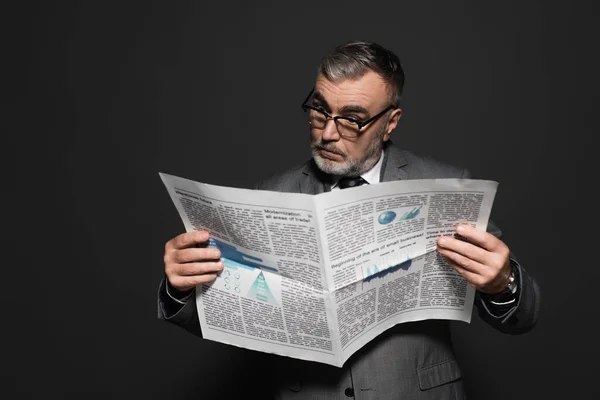 surprised businessman in eyeglasses reading newspaper isolated on dark grey