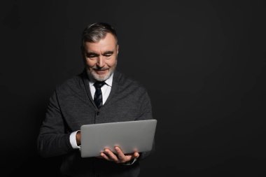 positive man in grey jumper and tie using laptop isolated on black