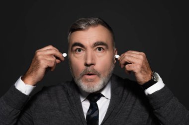 surprised senior man inserting earphones isolated on dark grey