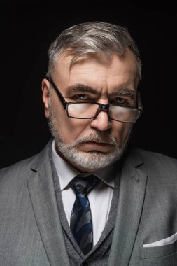 serious senior businessman in eyeglasses looking at camera isolated on black
