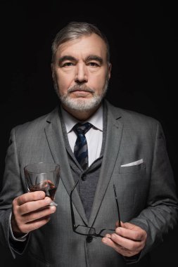 stylish senior man with glass of whiskey and eyeglasses looking at camera isolated on black