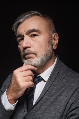 serious man in tie touching chin while looking at camera isolated on black