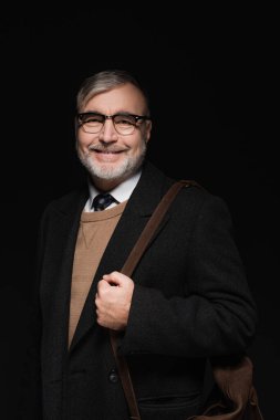 happy senior man in trendy coat and eyeglasses looking at camera isolated on black