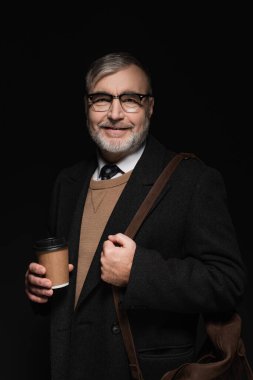 positive bearded man with leather bag and glass of whiskey looking at camera isolated on black