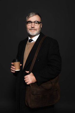 trendy senior man in coat holding takeaway drink and looking at camera on black