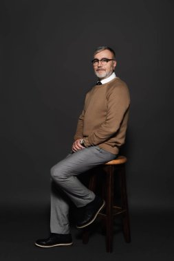 full length view of senior man in stylish clothes sitting on high stool on dark grey