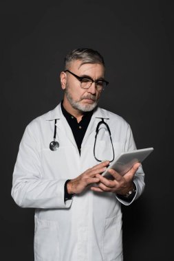 senior doctor in white coat and eyeglasses using digital tablet isolated on black clipart