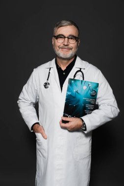 smiling physician in eyeglasses standing with magazine and hand in pocket of white coat isolated on dark grey clipart