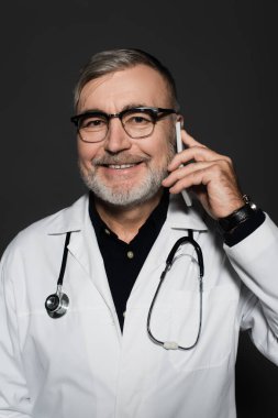 smiling senior doctor in eyeglasses talking on mobile phone isolated on dark grey clipart