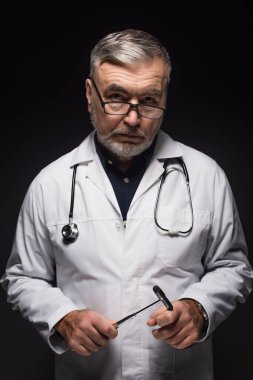 senior neurologist with reflex malleus and stethoscope looking at camera isolated on black