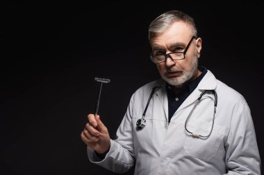 senior neurologist in eyeglasses holding reflex malleus and looking at camera isolated on black
