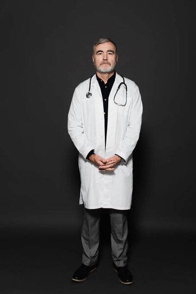 full length view of on senior physician in white coat looking at camera on dark grey