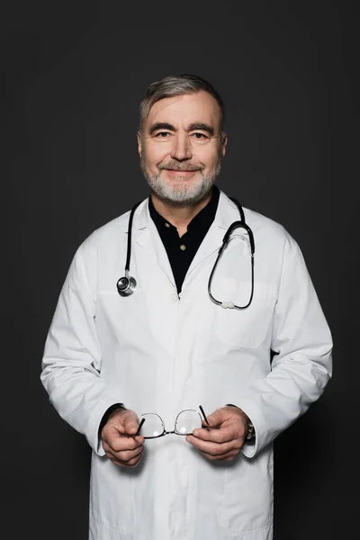 Positive Bearded Physician Holding Eyeglasses Smiling Camera Isolated Black — Foto de Stock